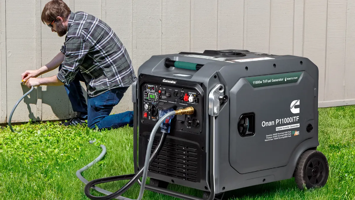 A Cummins Onan P11000iTF tri-fuel inverter generator running on the grass beside a house, its cord run to a connection point on the wall